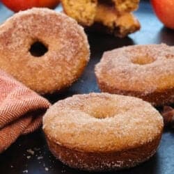 four apple cider donuts with a dusting of cinnamon sugar next to two apples and a star anise.