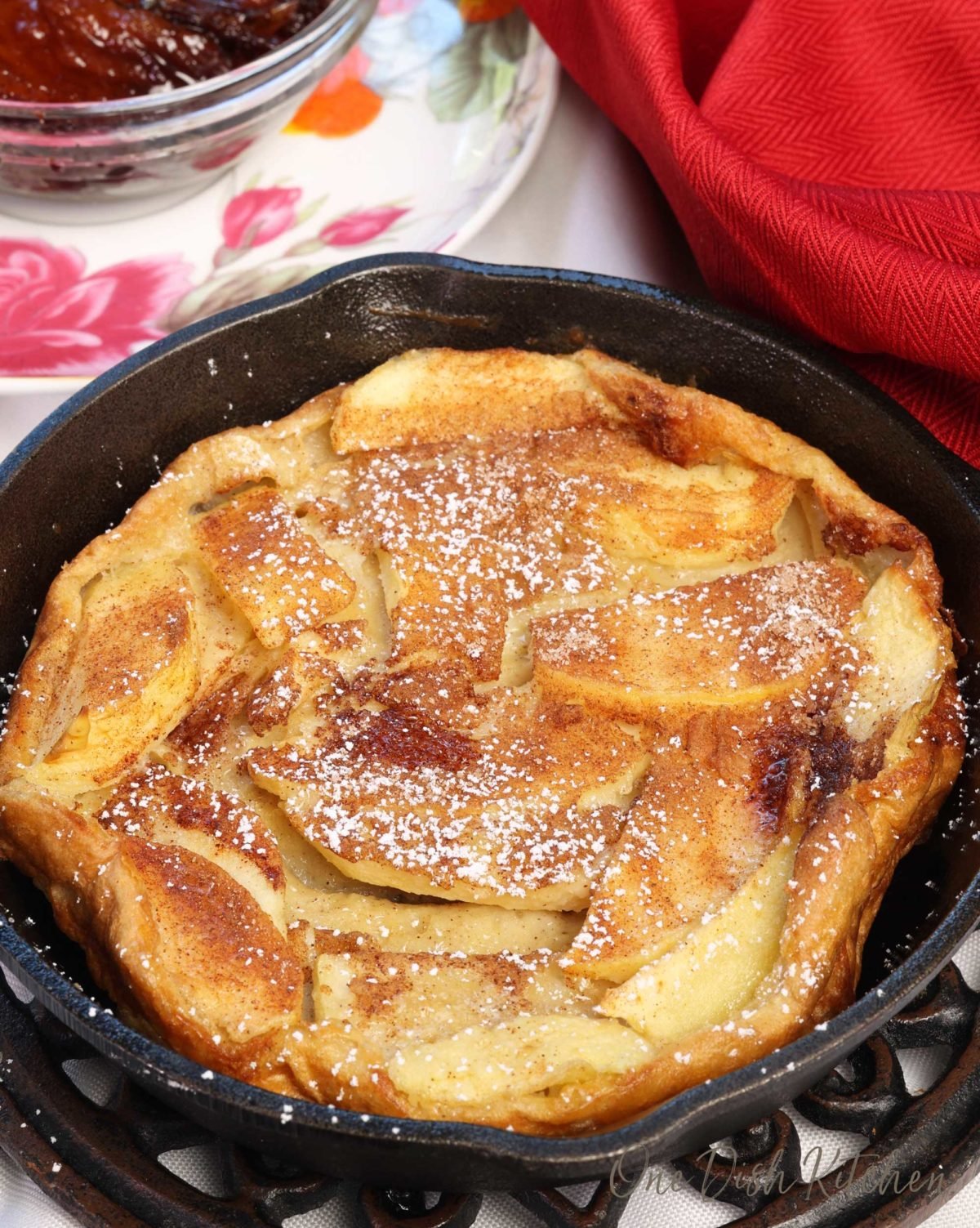 a mini apple dutch baby in a small cast iron skillet next to a red napkin and a bowl of fried apples.