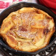 a mini apple dutch baby in a small cast iron skillet next to a red napkin and a bowl of fried apples.