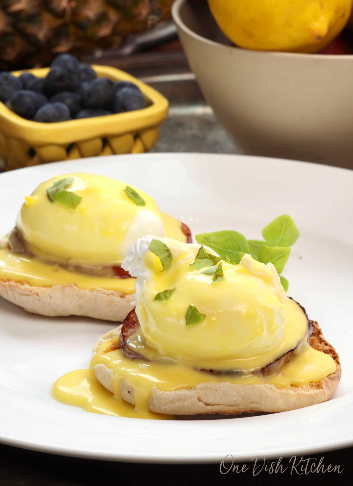 two eggs benedict with hollandaise sauce over the top on a white plate next to a yellow bowl of blueberries.