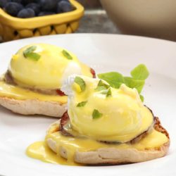 two eggs benedict with hollandaise sauce over the top on a white plate next to a yellow bowl of blueberries