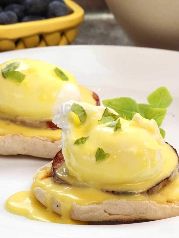two eggs benedict with hollandaise sauce over the top on a white plate next to a yellow bowl of blueberries