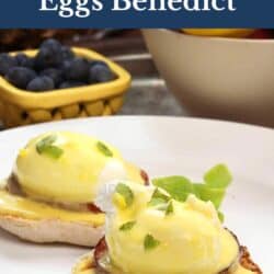 two eggs benedict on a white plate next to a bowl of blueberries.