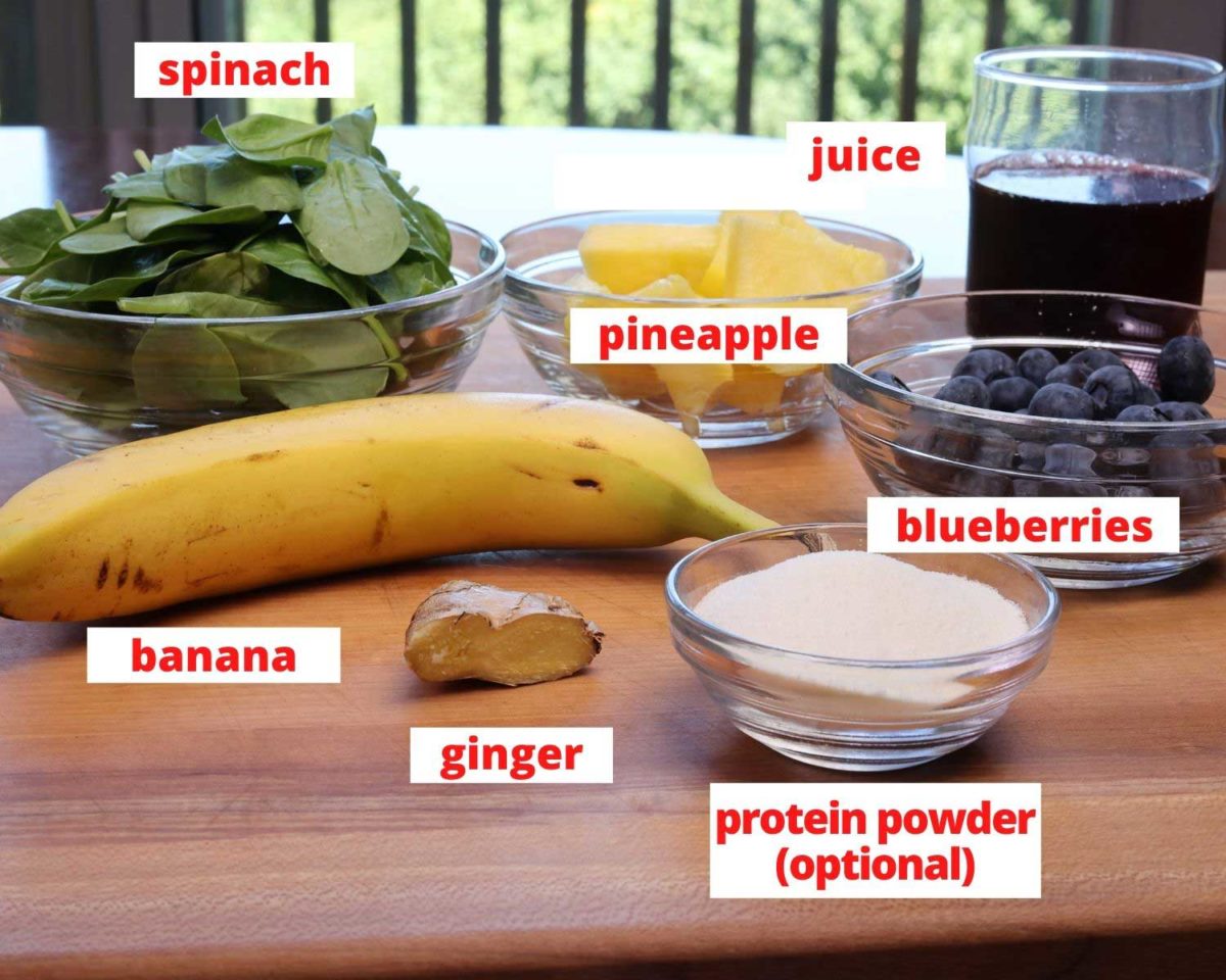 spinach, a bowl of blueberries, pineapple, a banana, and a knob of ginger on a wooden cutting board in a kitchen.
