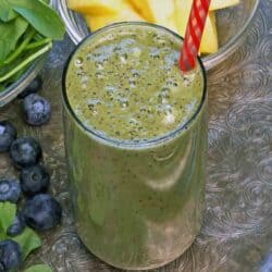 a green smoothie on a silver tray next to a bowl of fresh spinach, blueberries and pineapple.