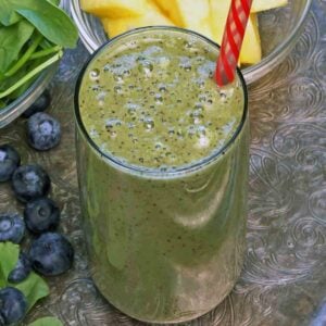 a green smoothie on a silver tray next to a bowl of fresh spinach, blueberries and pineapple.