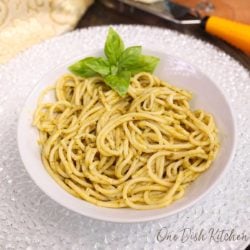 a white bowl filled with pesto pasta with a wedge of parmesan on the side of the table