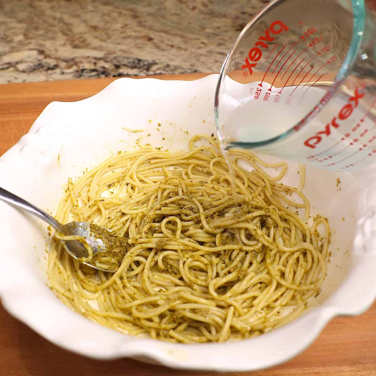 pesto water in a small measuring cup being poured over pesto pasta.