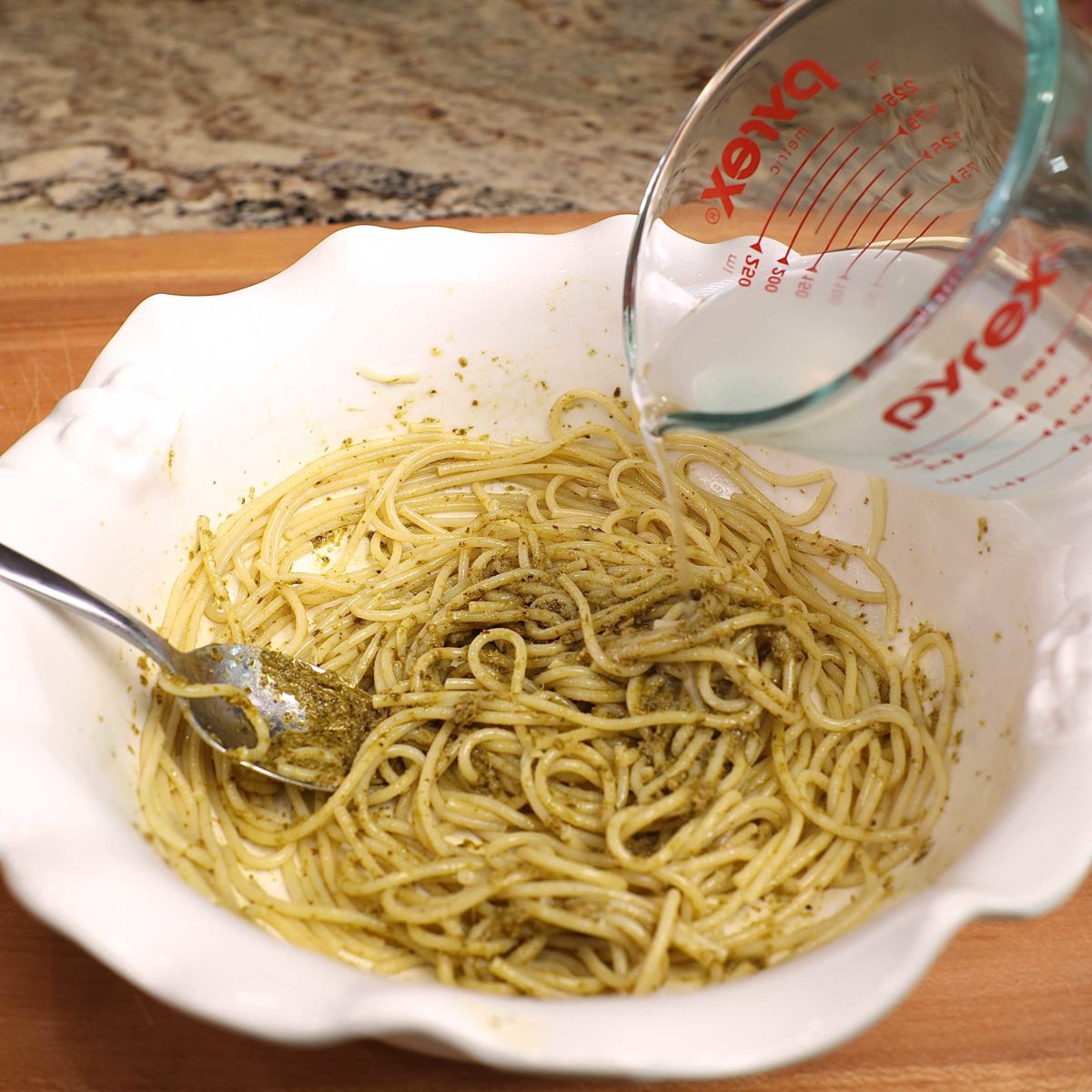 pesto water in a small measuring cup being poured over pesto pasta.