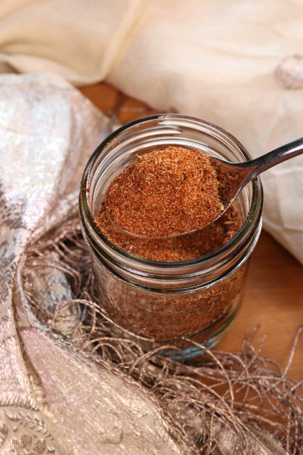 a spoonful of taco seasoning held over an open jar of the seasoning next to a cream colored lace scarf