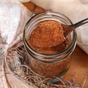 a spoonful of taco seasoning held over an open jar of the seasoning next to a cream colored lace scarf