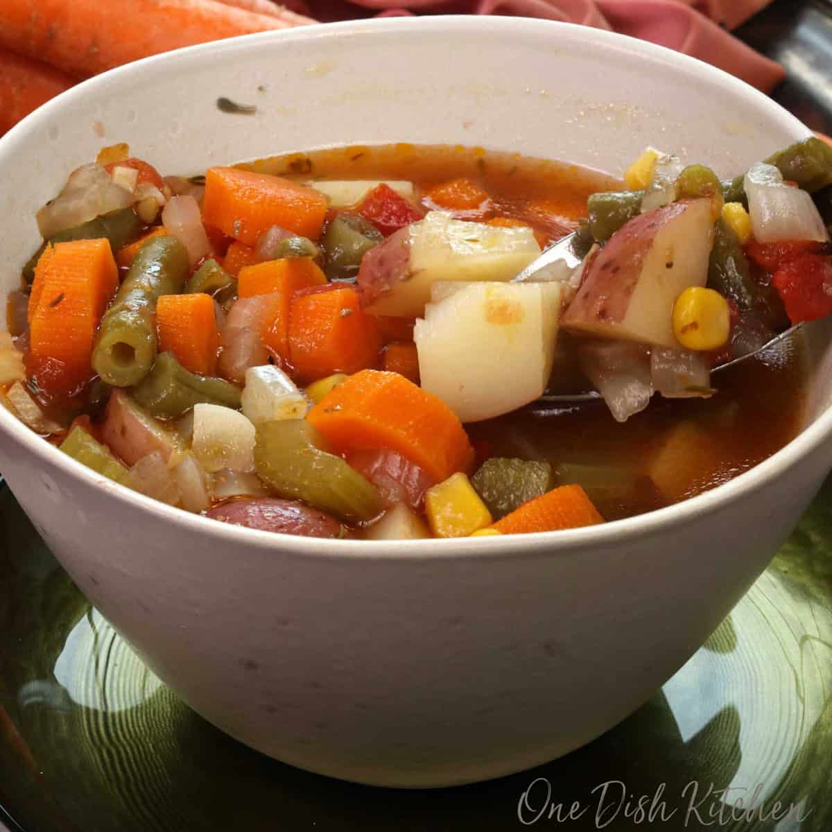 a bowl of vegetable soup with a spoon on the side next to two carrots and an orange napkin.