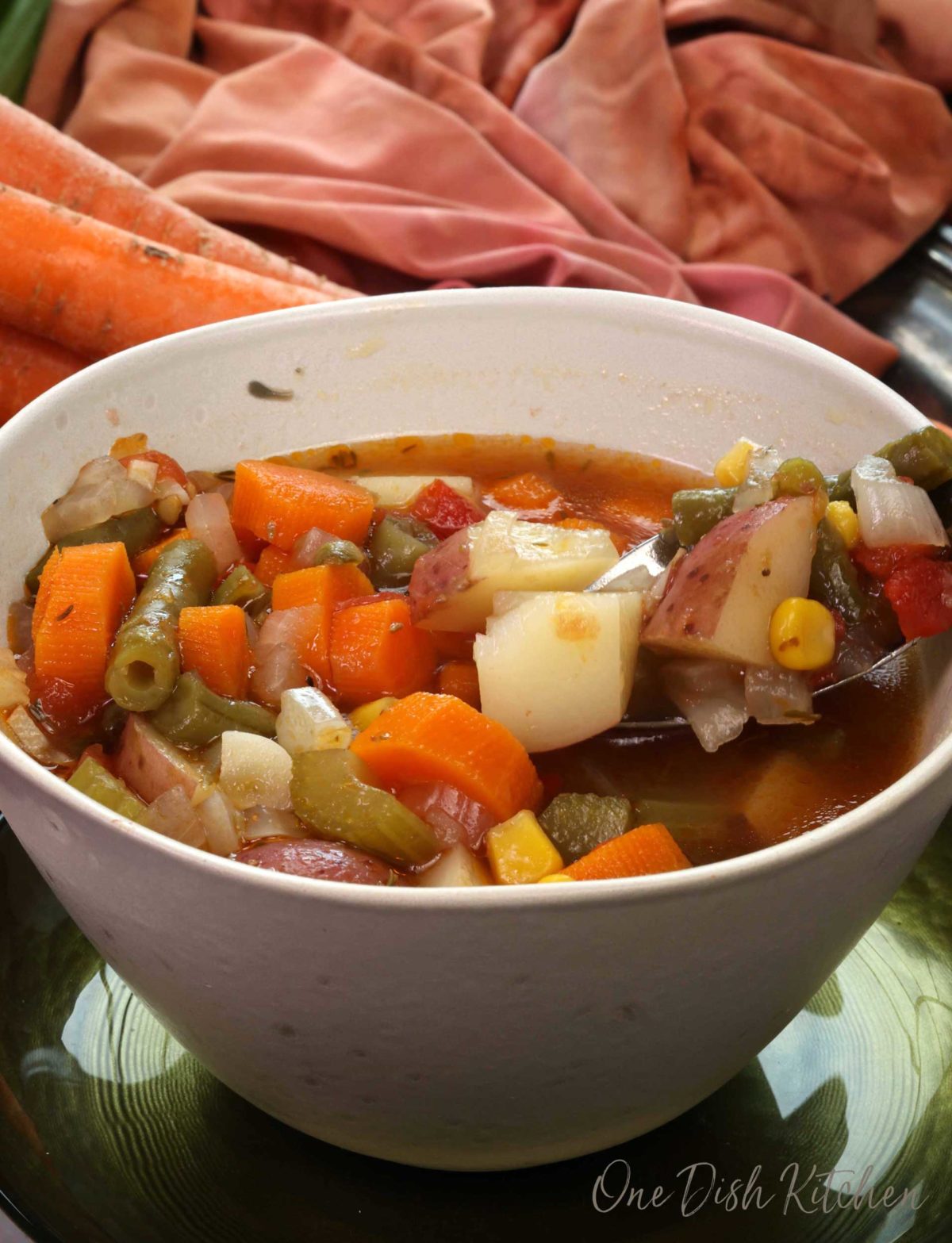 a bowl of vegetable soup with a spoon on the side next to two carrots and an orange napkin.