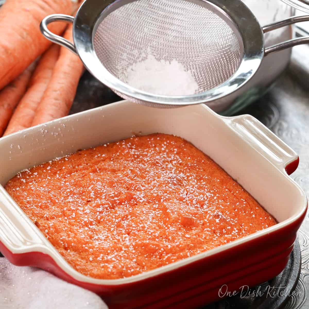 a small baked carrot souffle in a square red baking dish next to a container of powdered sugar and raw carrots.