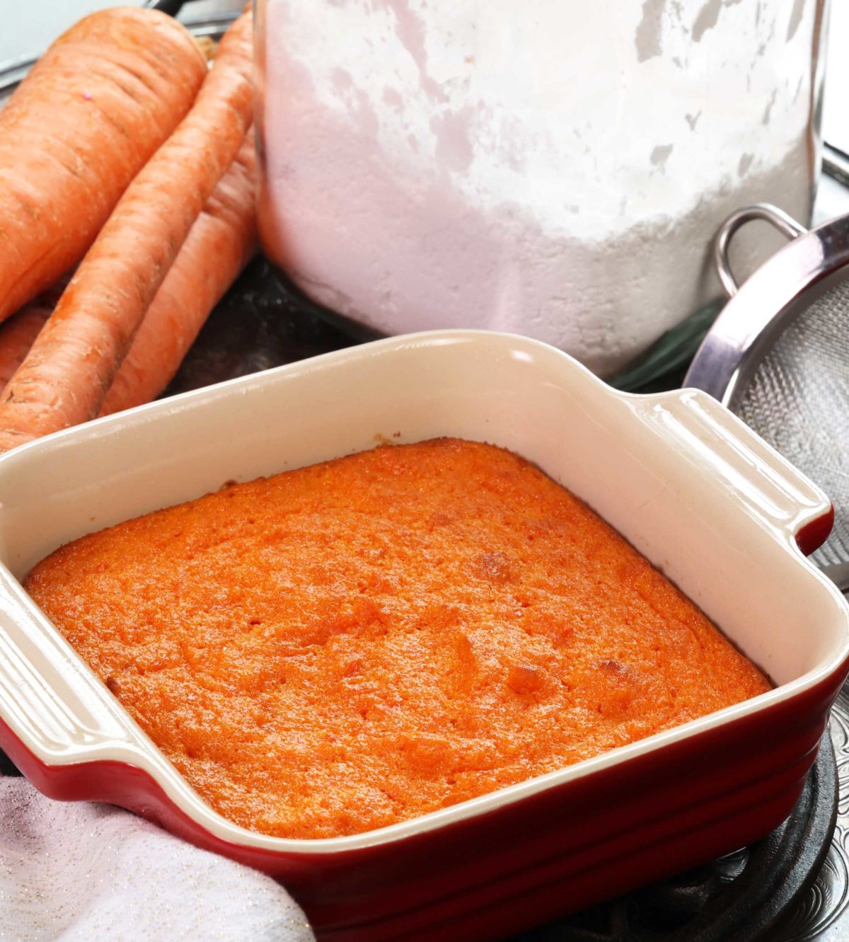 a small baked carrot souffle in a square red baking dish next to a container of powdered sugar and raw carrots.