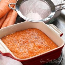 a small baked carrot souffle in a square red baking dish next to a container of powdered sugar and raw carrots