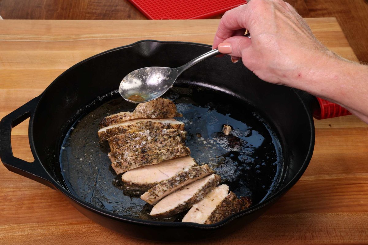 sliced turkey tenderloin medallions in a cast iron skillet with a spoon overhead drizzling the butter sauce over the tops of the pieces.