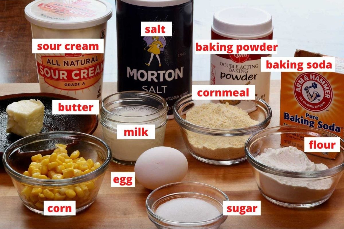 cornmeal, an egg, baking powder, corn, salt and other ingredients needed to make corn muffins on a cutting board.