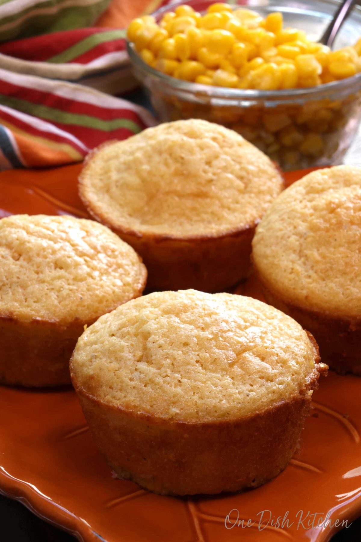 four corn muffins on a plate next to a bowl of corn.