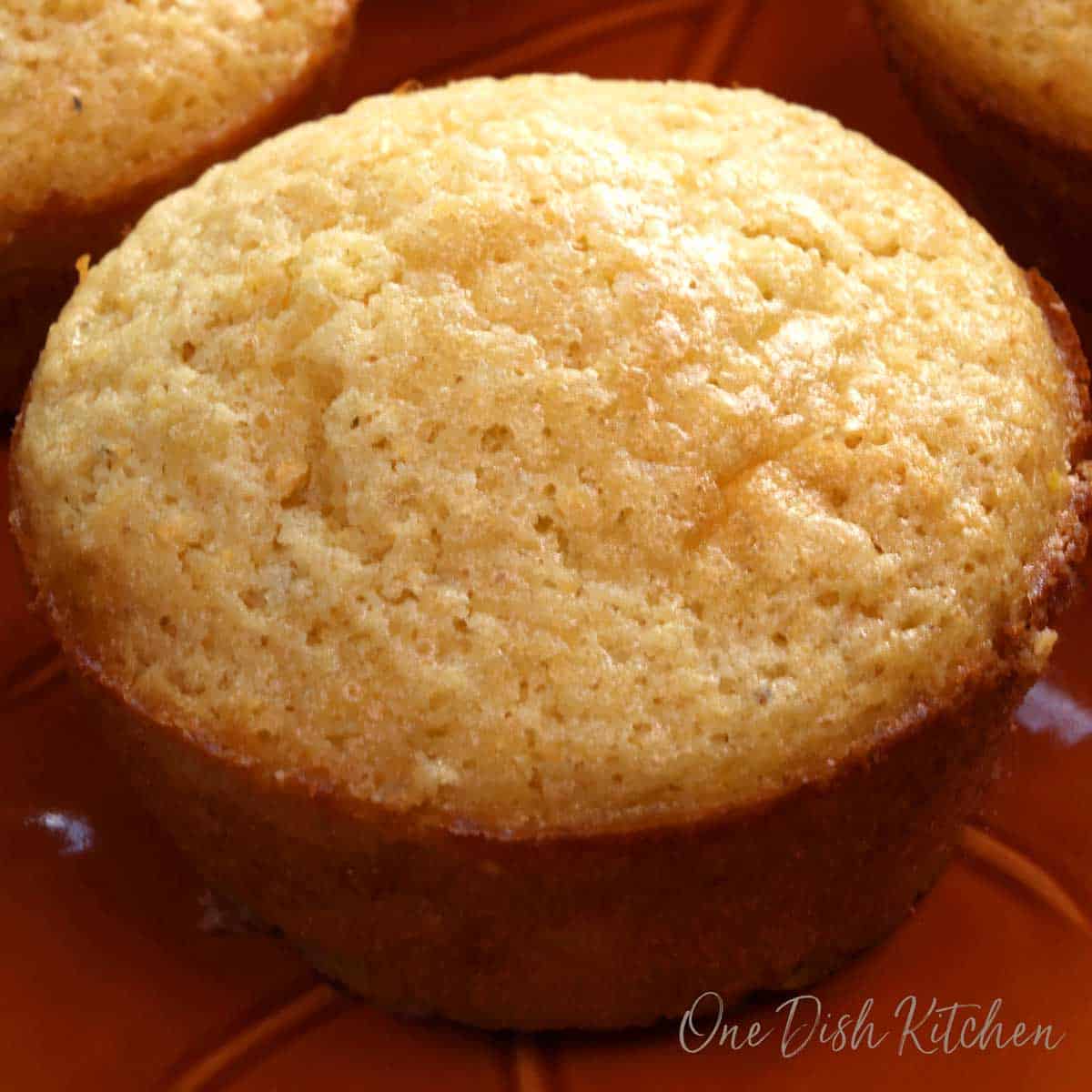 a baked corn muffin on an orange plate next to three other muffins.