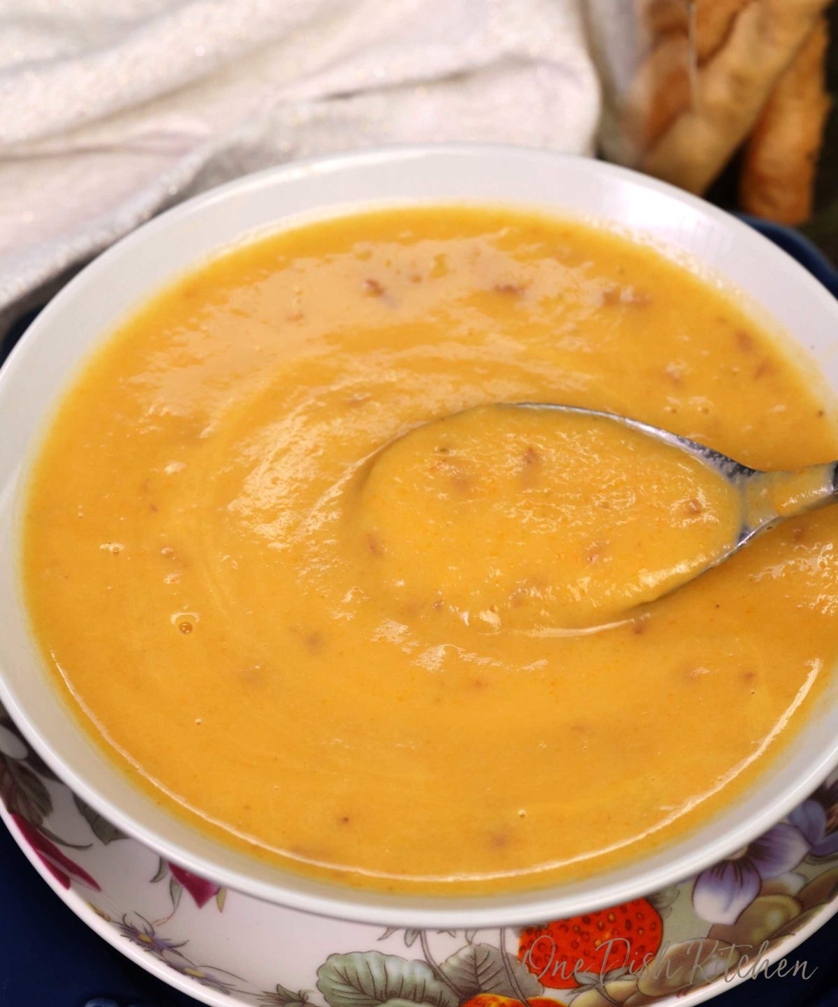 a spoon in a bowl of creamy vegetable soup next to a jar of breadsticks.