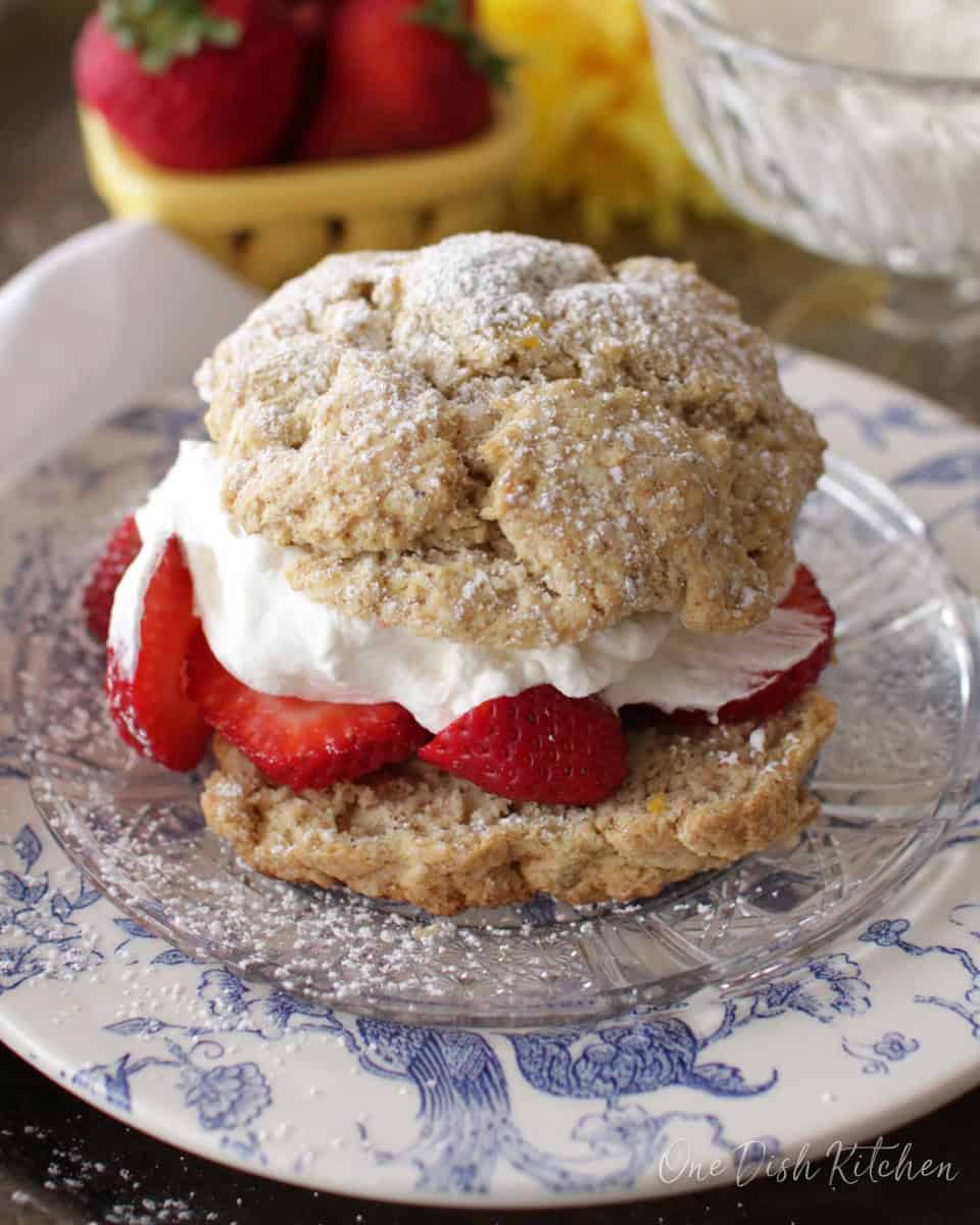 a single strawberry shortcake with whipped cream on a white and blue plate with strawberries in a bowl in the background.