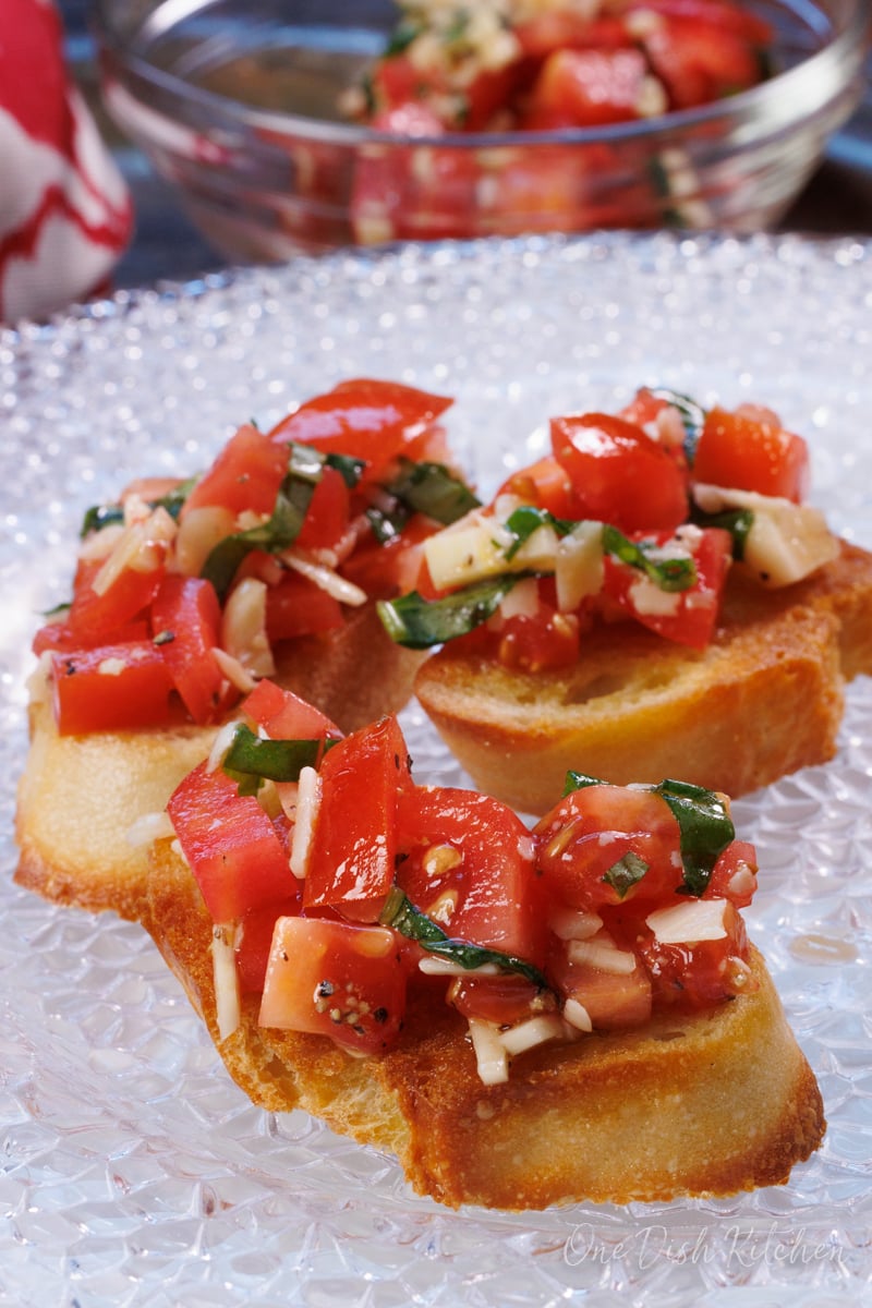 a piece of tomato and basil bruschetta on a white plate next to a bowl of chopped tomatoes.