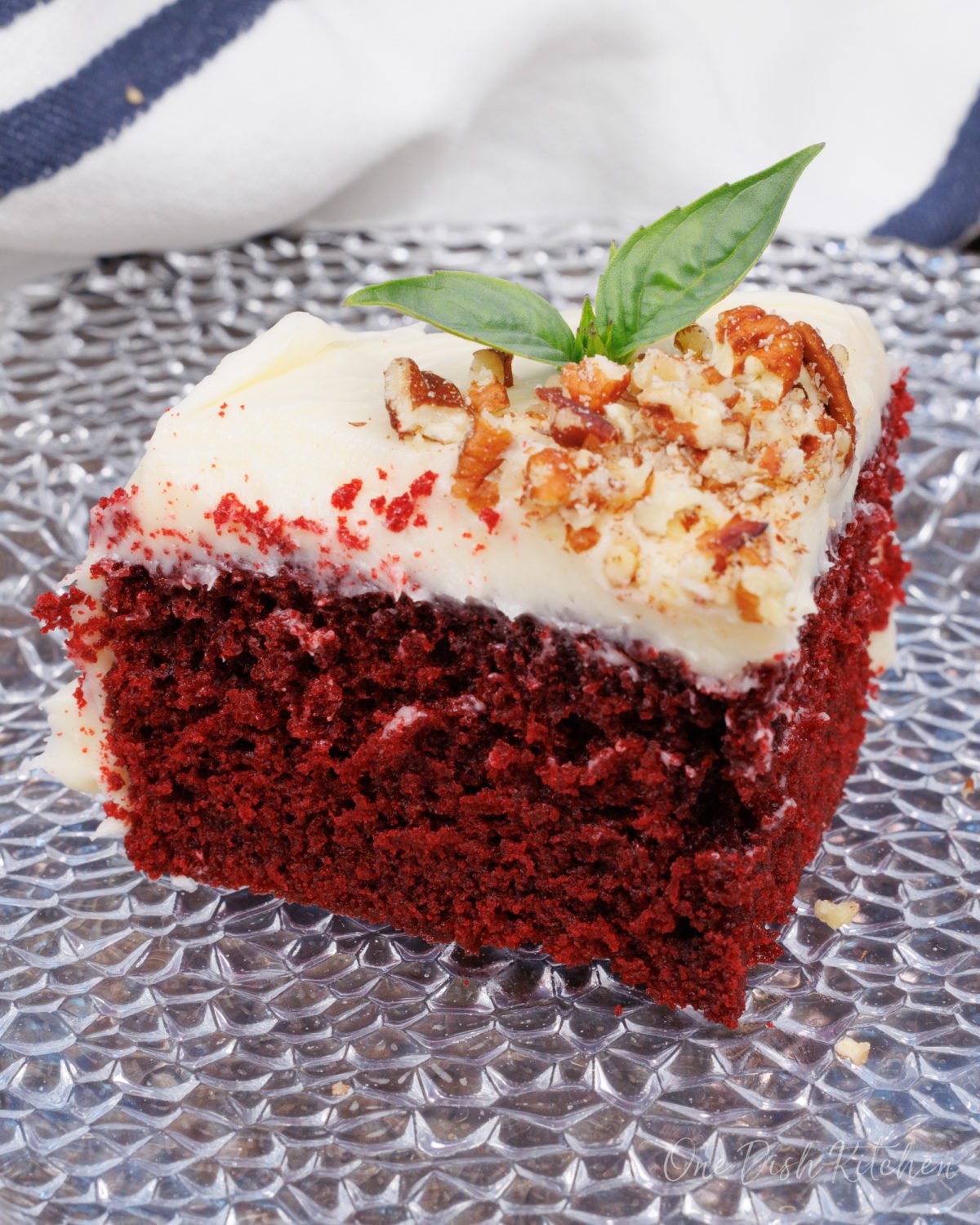 a slice of red velvet cake with cream cheese frosting and a sprig of fresh mint on a clear plate.