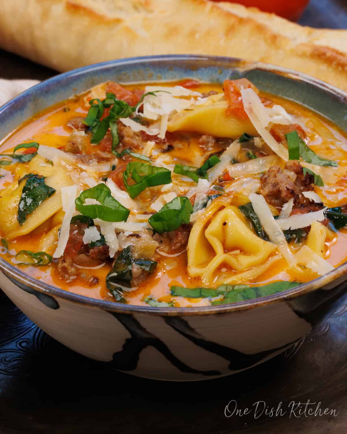 a bowl of tortellini soup on a silver tray next to a loaf of french bread and two tomatoes.