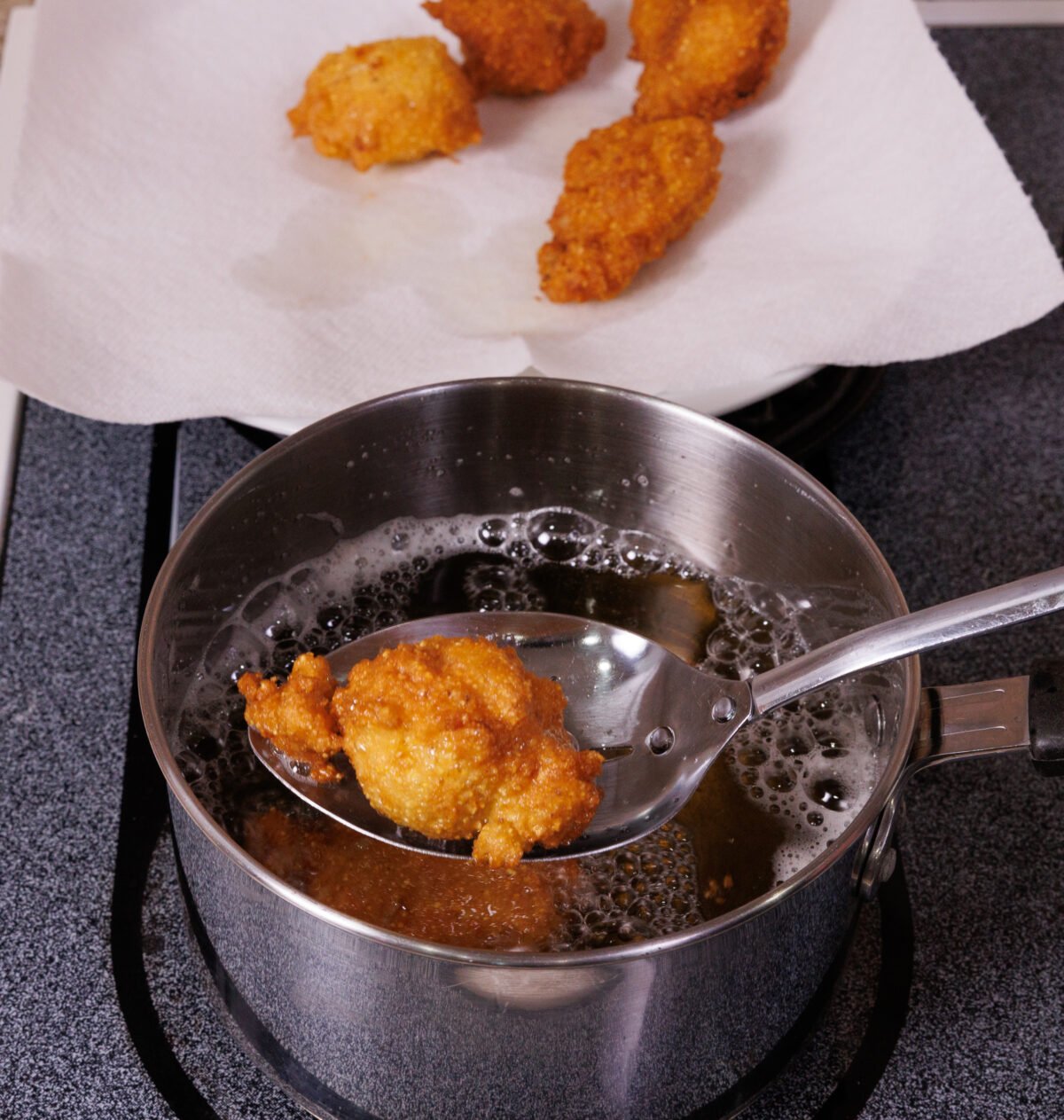 removing fried hushpuppies from a saucepan with a paper towel lined plate in the background.