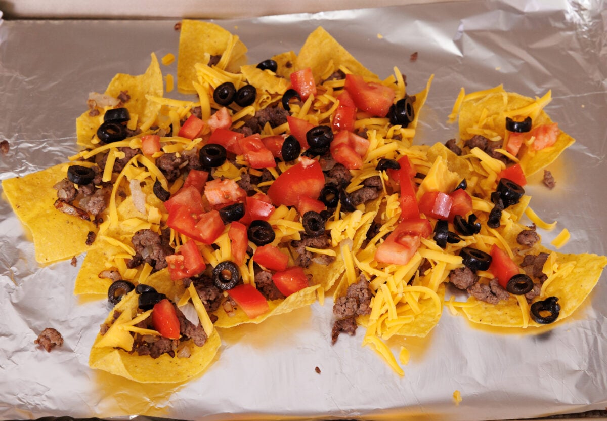 tortilla chips topped with ground beef, cheese, tomatoes, and olives on a baking sheet.