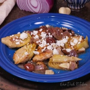 single serving of Mediterranean chicken on a blue plate surrounded by a red onion and a bowl of artichoke hearts.