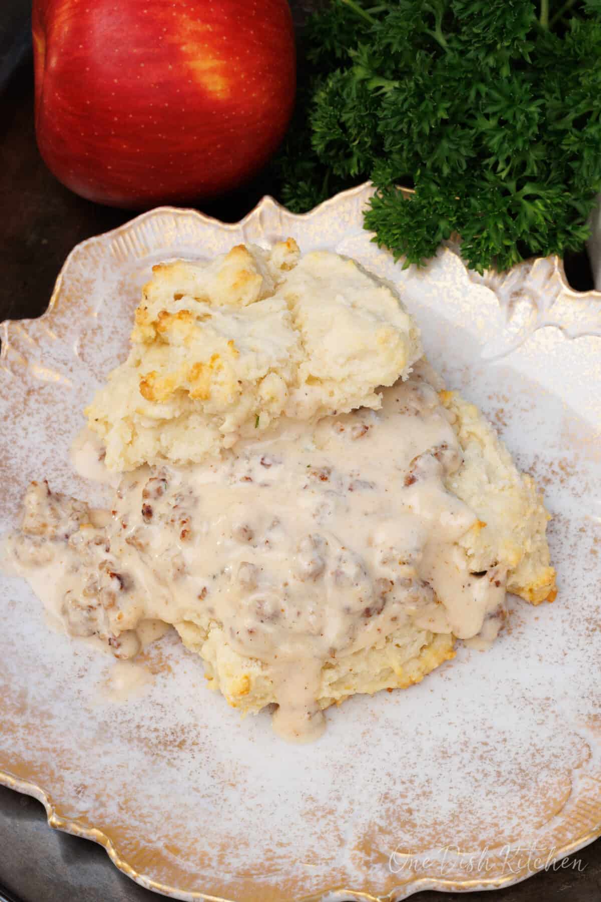 one biscuit with sausage gravy on a white plate next to an apple.