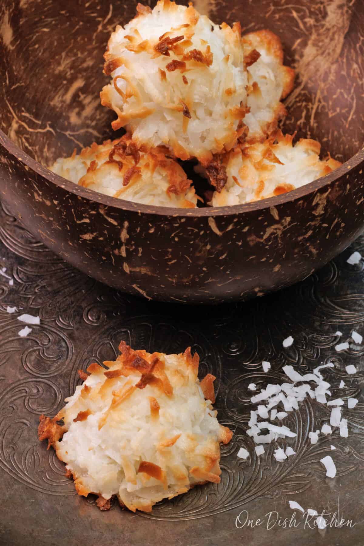 coconut macaroons on a silver try next to coconut flakes scattered around the cookie.