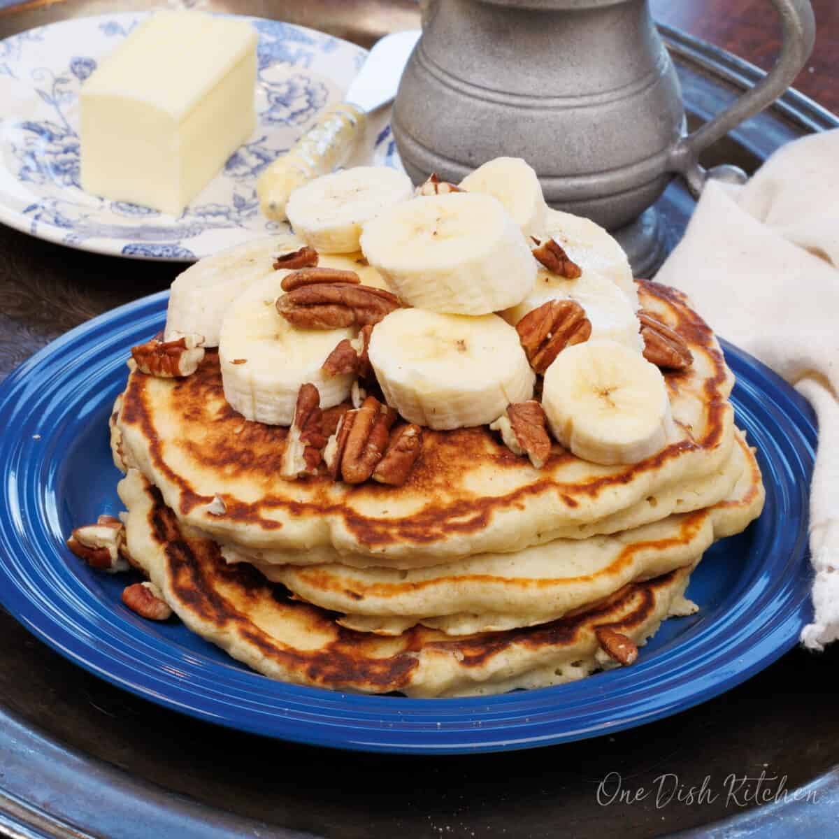 banana pancakes on a plate next to a plate of butter and a jug of syrup.
