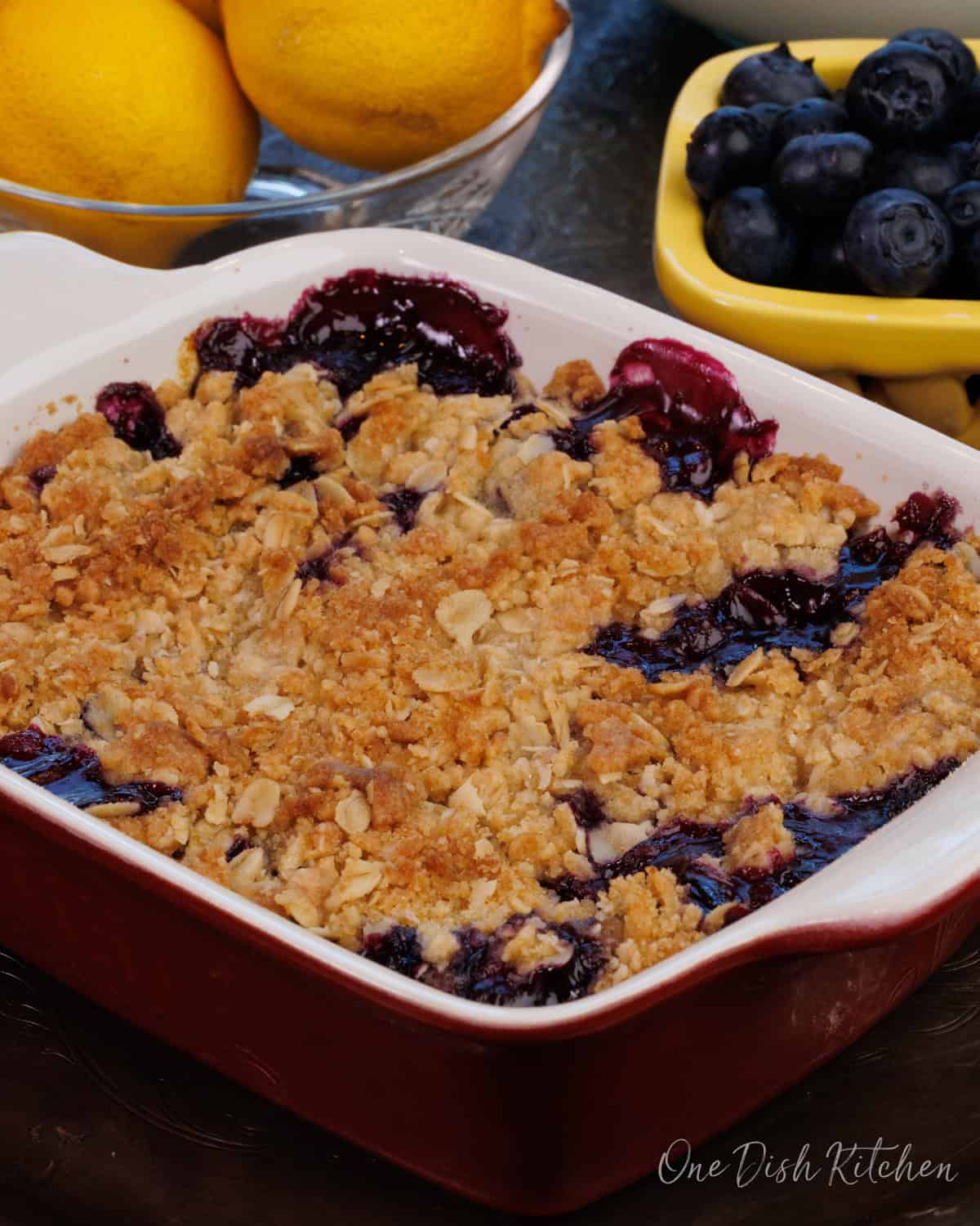 a blueberry crisp in a rectangular baking dish next to a bowl of blueberries.