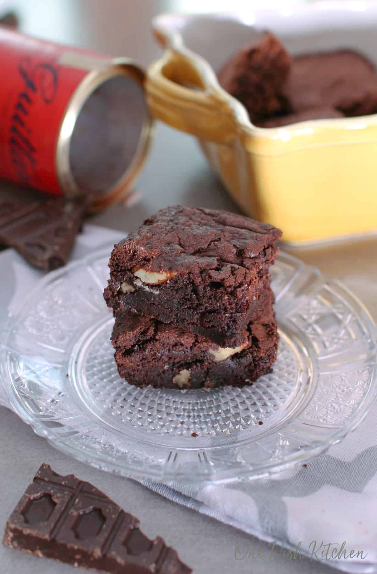 two brownies on top of each other on a white plate next to a bar of chocolate and a container of cocoa powder.