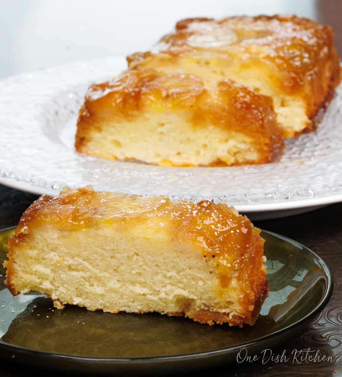 a slice of banana upside down cake on a green plate next to the cake loaf on another large plate.