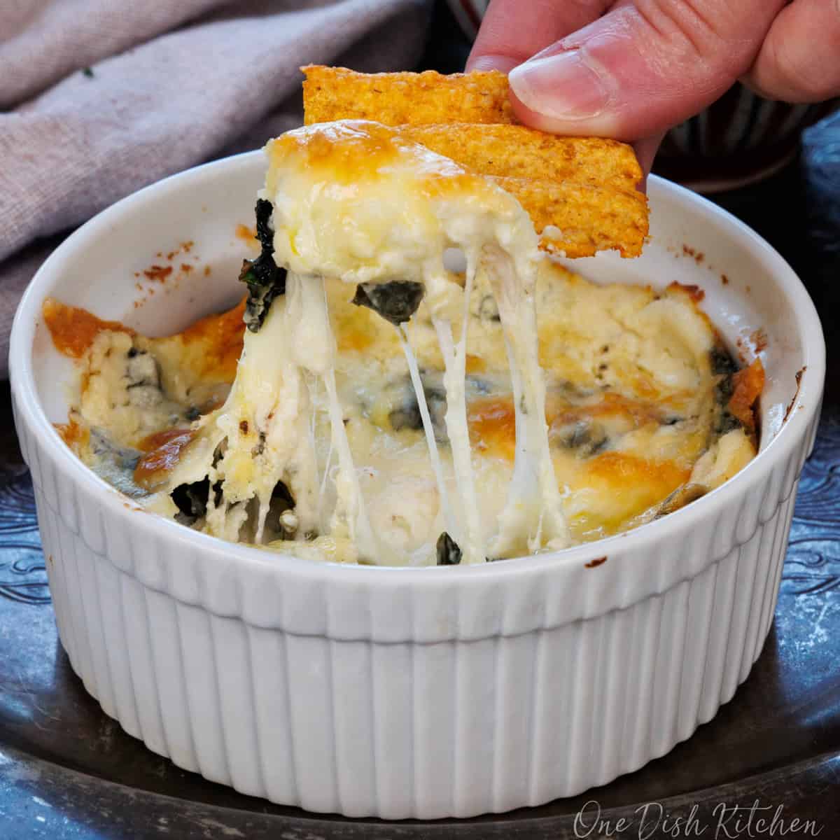 a single serving hot spinach dip in a ramekin with a chip on the side.