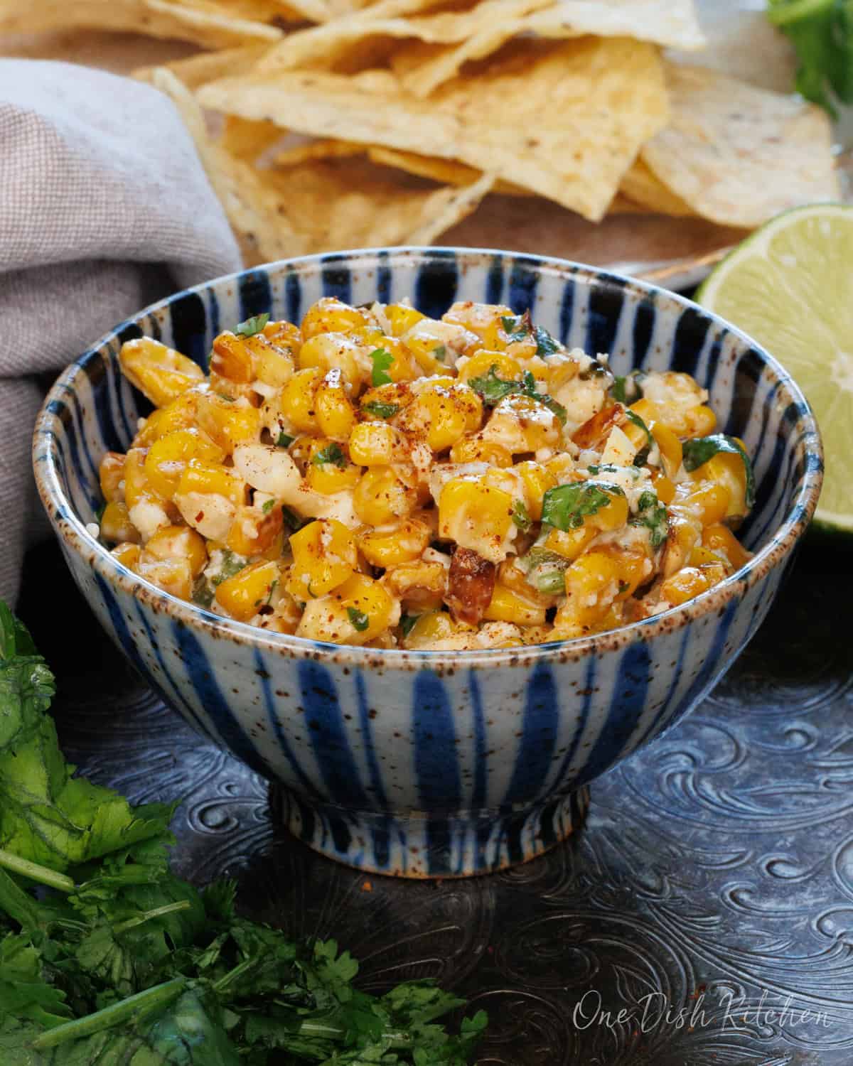 a bowl of esquites next to a lime and tortilla chips on a silver tray.