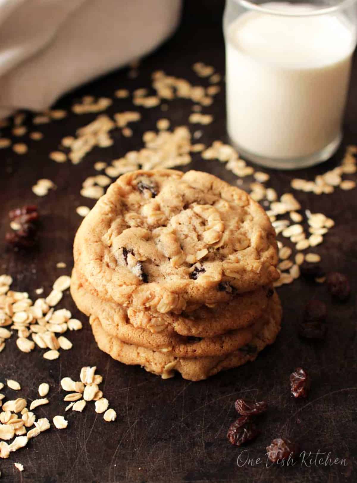 a stack of 3 oatmeal raisin cookies on a black tray surrounded by raisins.