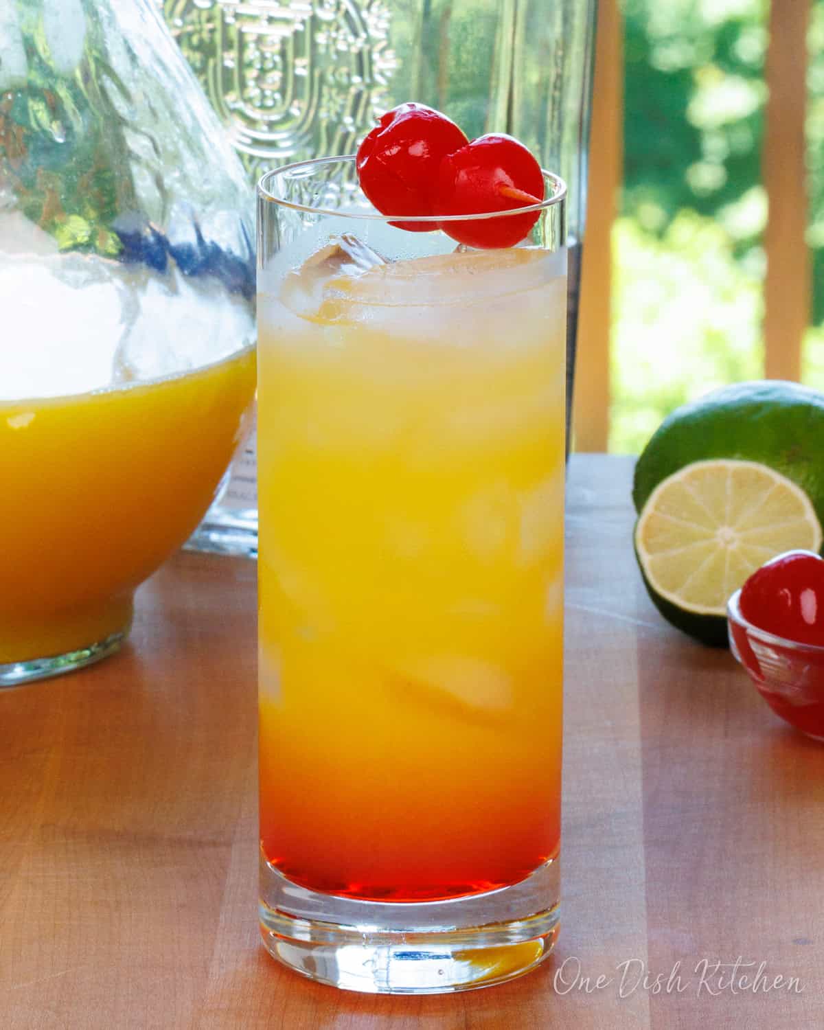 a tequila sunrise cocktail on a wooden table next to a bowl of cherries and a lime.