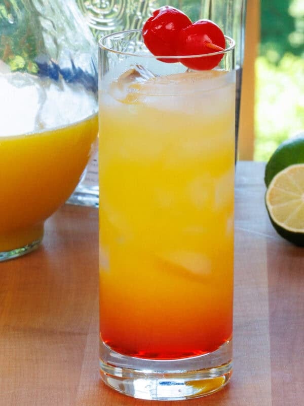 a tequila sunrise cocktail on a wooden table next to a bowl of cherries and a lime.