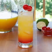 a tequila sunrise cocktail on a wooden table next to a bowl of cherries and a lime