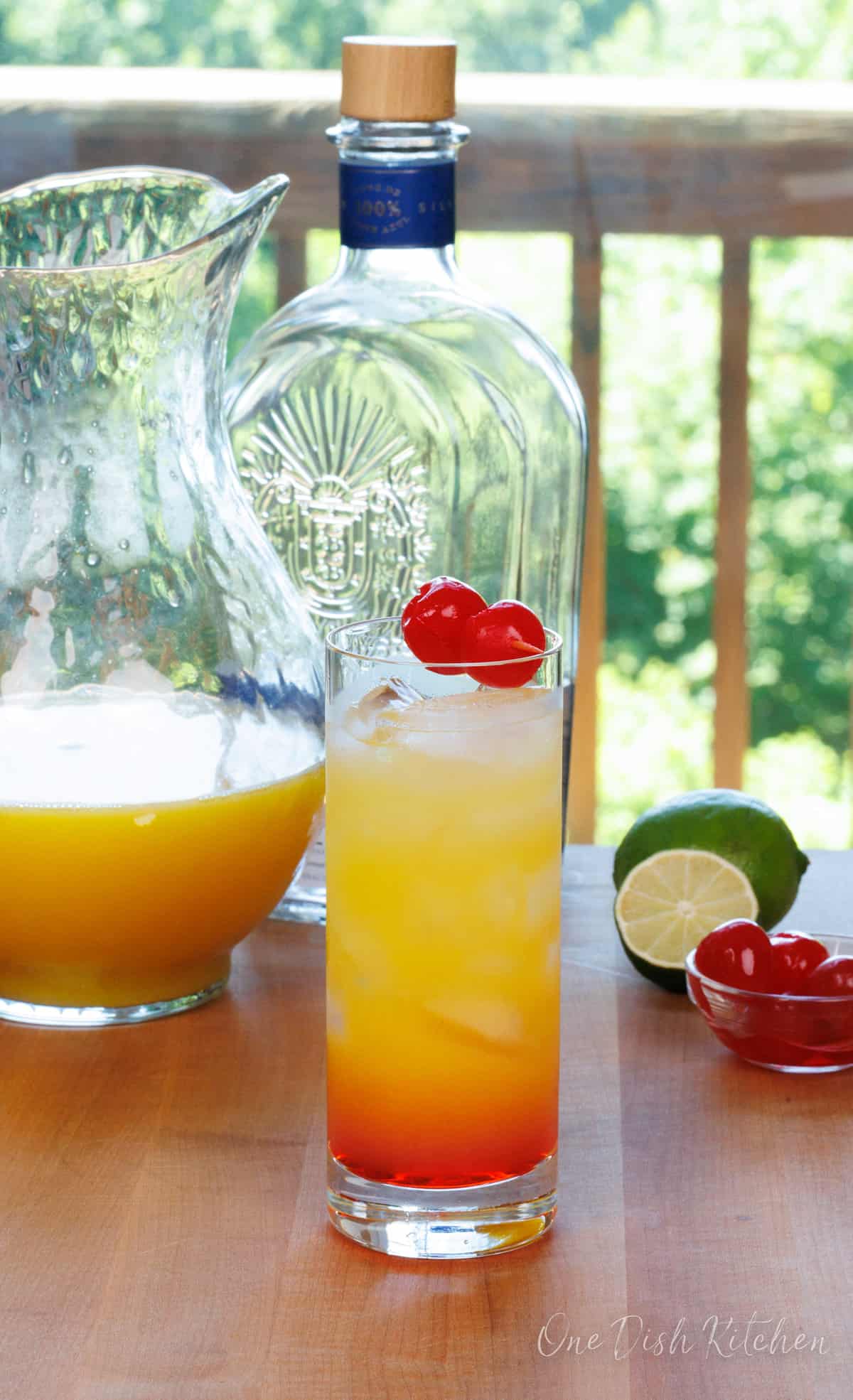 a tequila sunrise topped with maraschino cherries next to a pitcher of orange juice