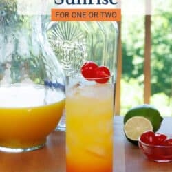 a tequila sunrise on a kitchen table next to a pitcher of orange juice and a lime.