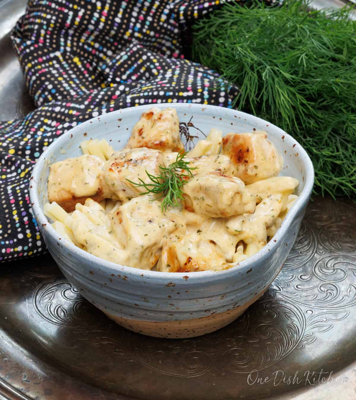 a blue bowl filled with creamy mustard chicken on a silver tray next to fresh dill