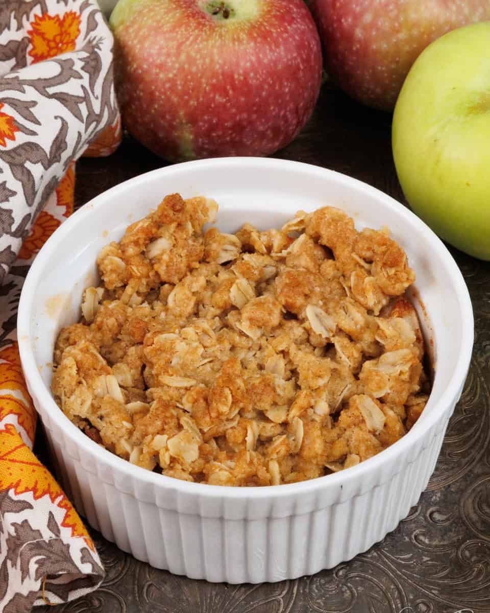 an apple crisp on a silver tray next to 5 apples and a bowl of whipped cream.