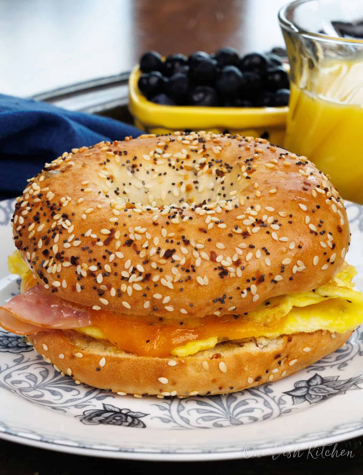 a breakfast bagel with ham and cheese on a blue plate next to a glass of orange juice.