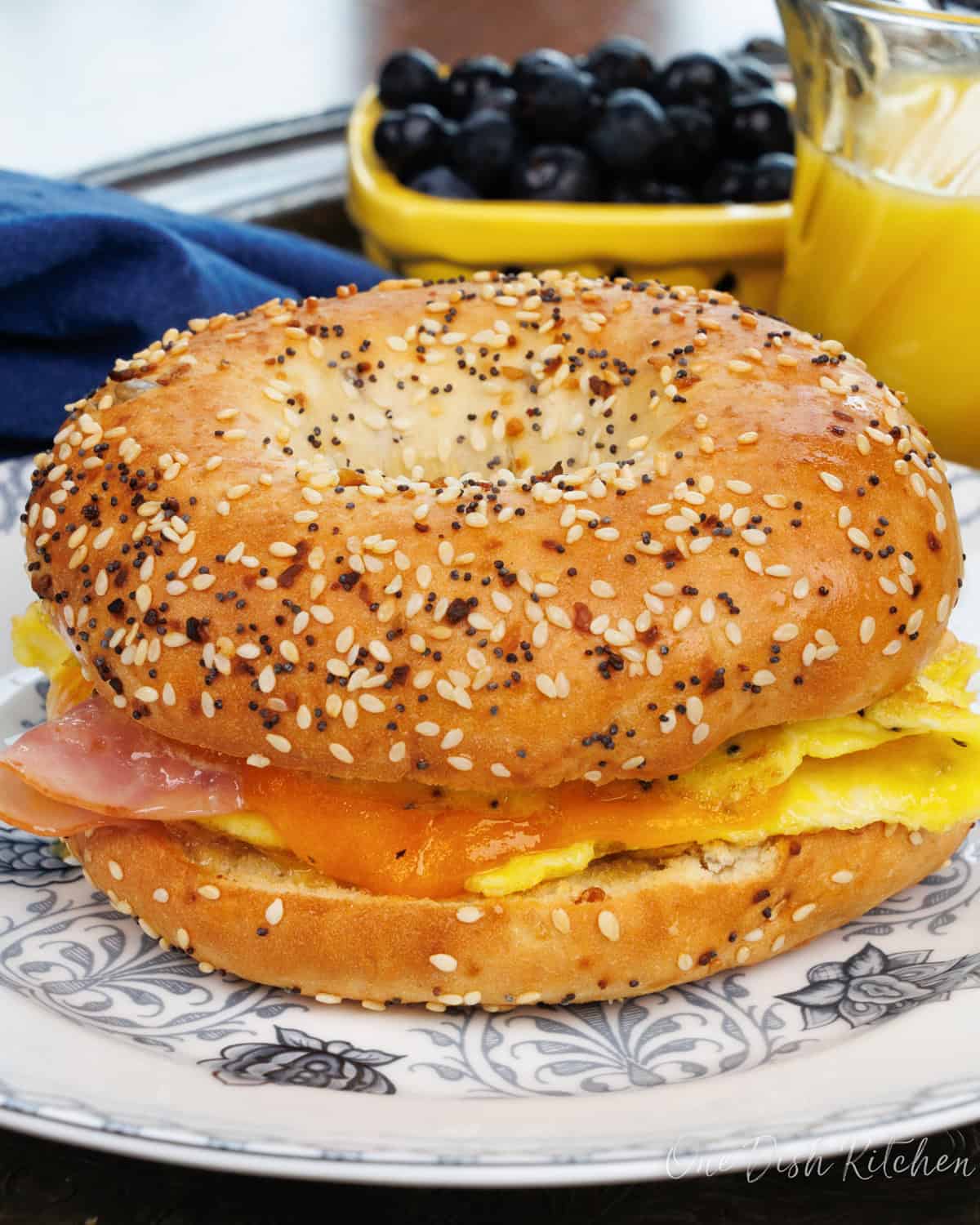 a breakfast bagel with ham and cheese on a blue plate next to a glass of orange juice.